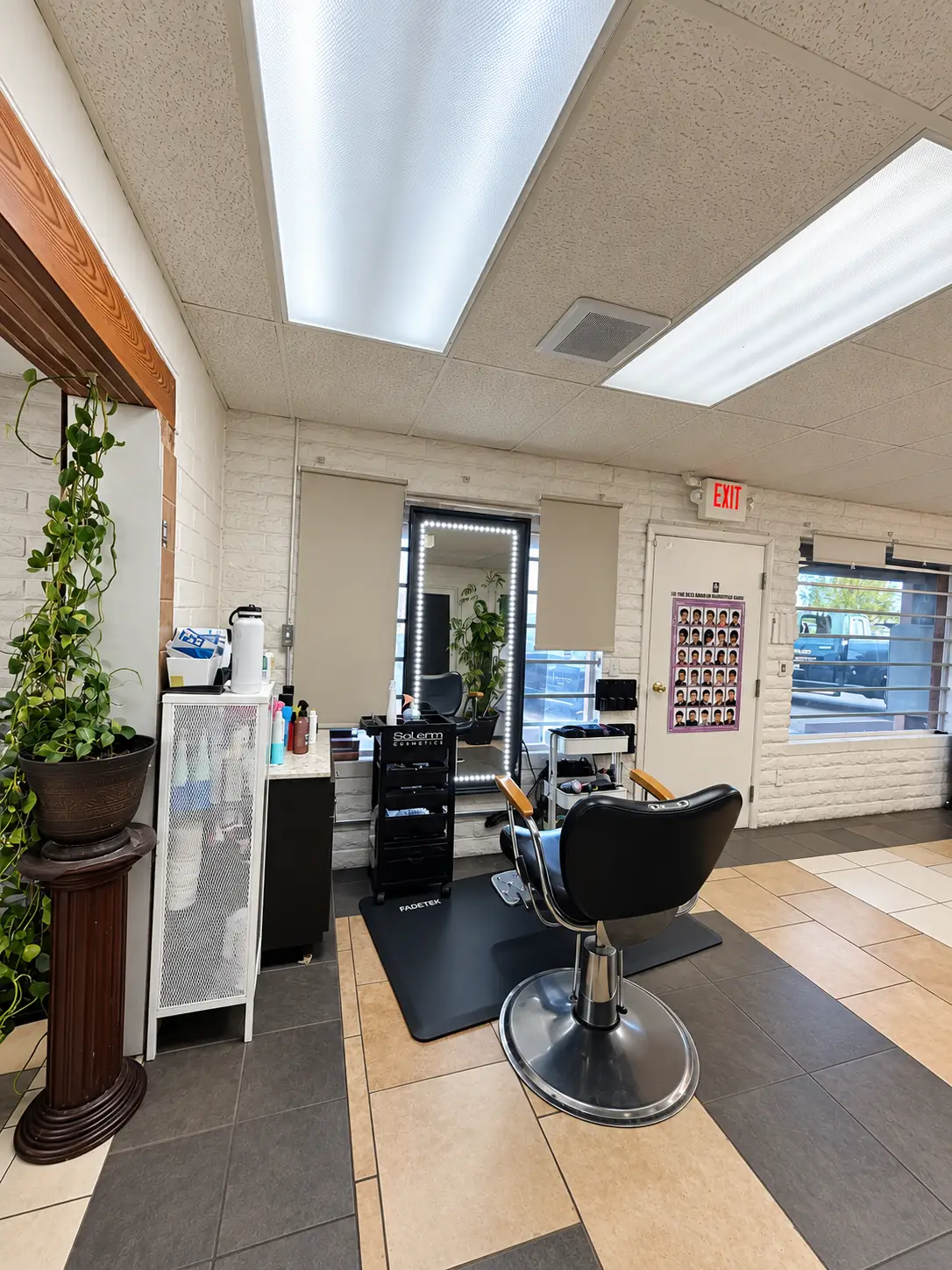 A black styling chair on a mat beside a white brick wall, a tall plant and a lit mirror