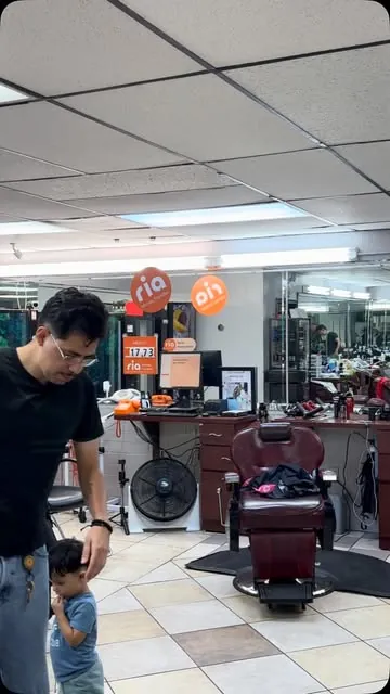 Inside the barbershop: a barber in glasses stands near a burgundy chair while a small child walks beside him