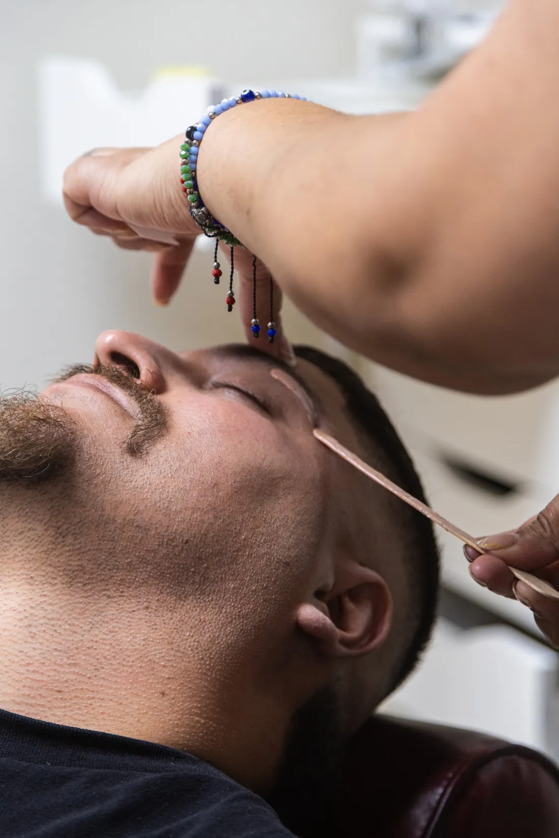 A client lies back with eyes closed while wax is applied to his eyebrow with a wooden spatula