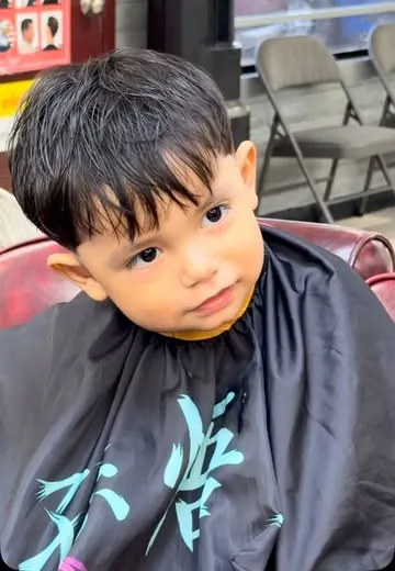 A toddler in a black cape sits calmly in a barber chair during his haircut