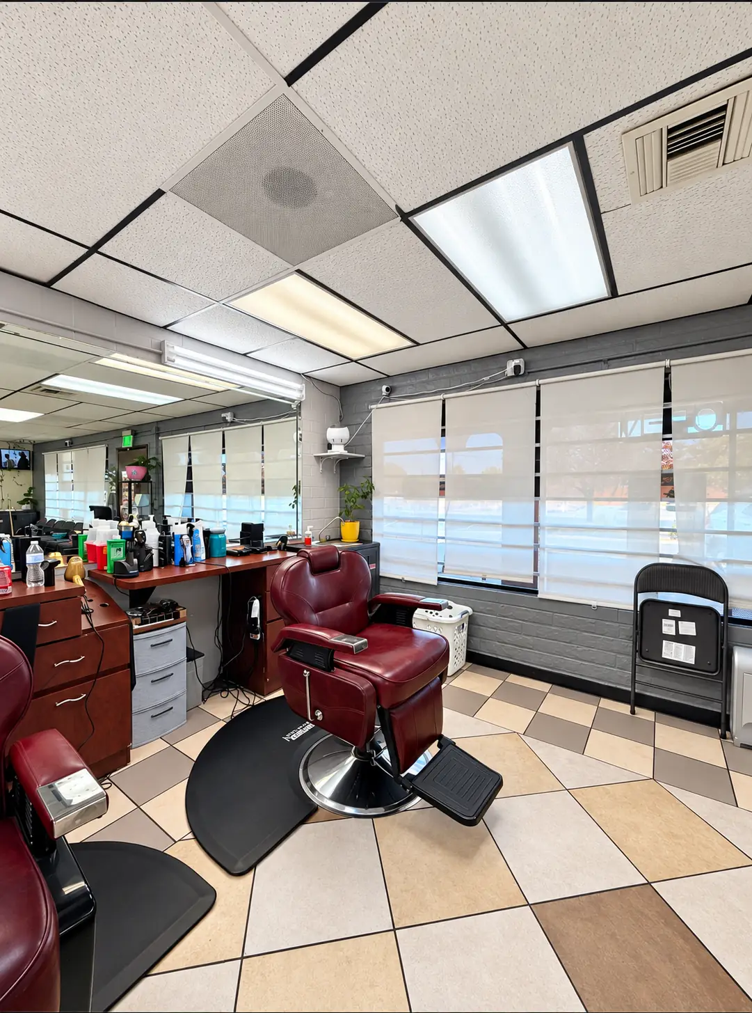 A burgundy barber chair by the front windows with shades, next to a station counter and mirror
