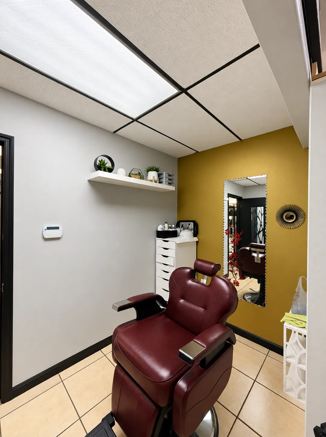 A burgundy leather barber chair in a station with a gold accent wall, mirror and tile floor at Ema's Family Barbershop